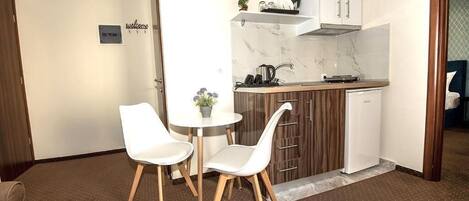 Una cocina compacta con armarios blancos, salpicadero de mármol, un pequeño comedor con dos sillas blancas y una planta en maceta.