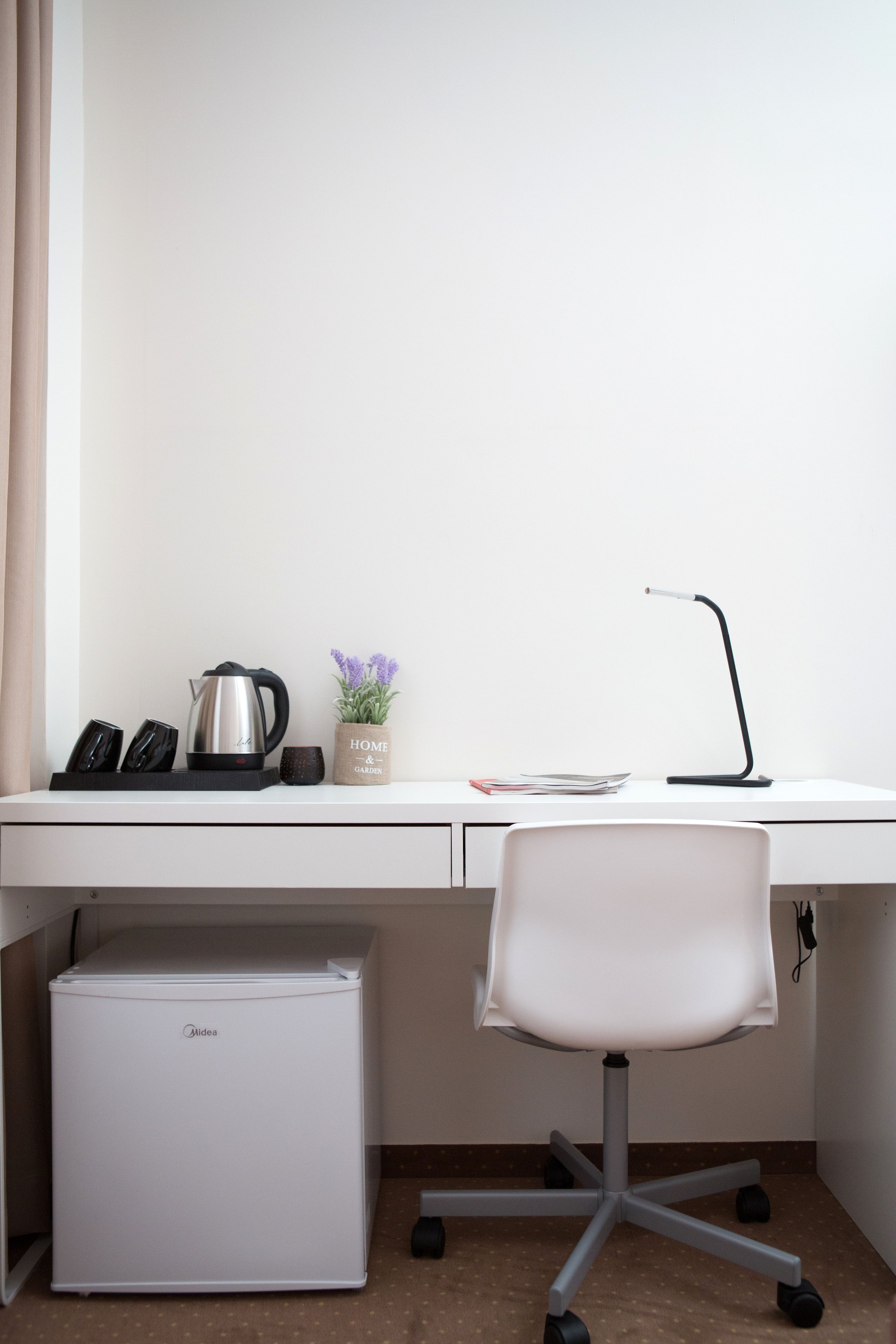 Een wit bureau met een waterkoker, een bloempot, een lamp en een witte stoel.