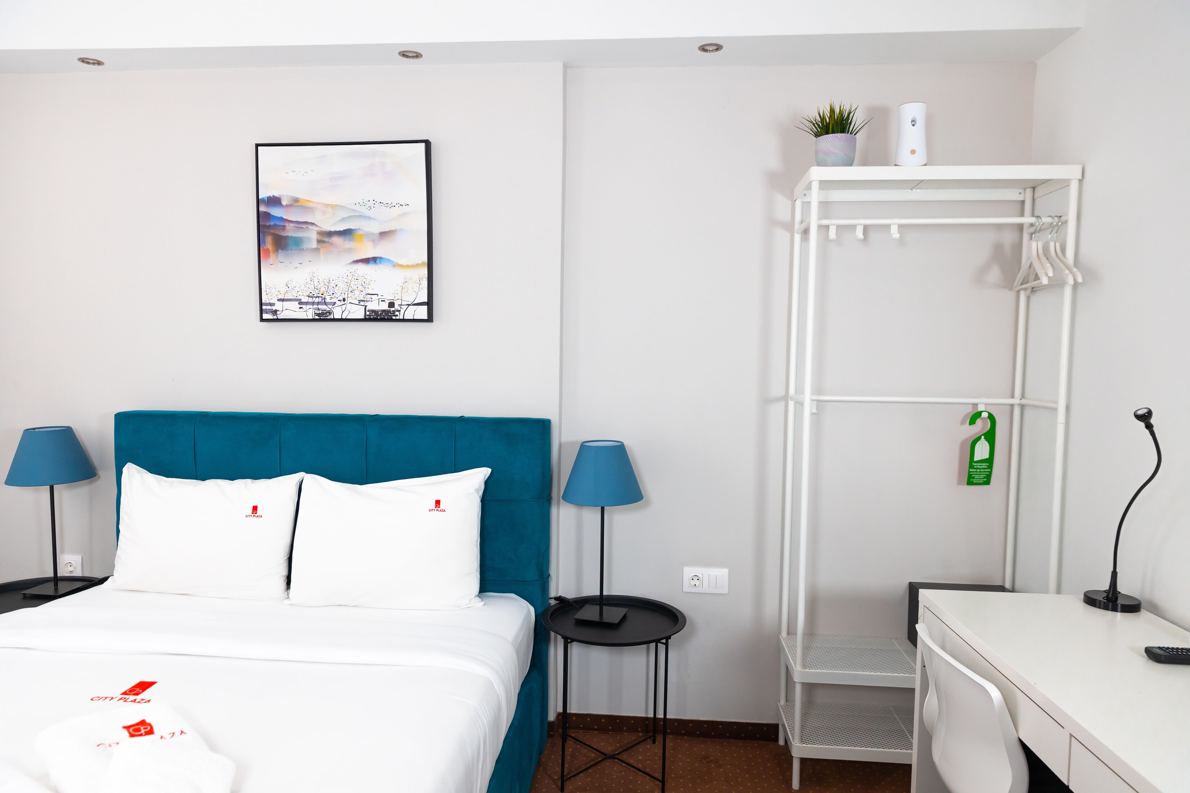 A hotel room with a bed, two lamps, a desk, a white wardrobe, and a framed picture on the wall.