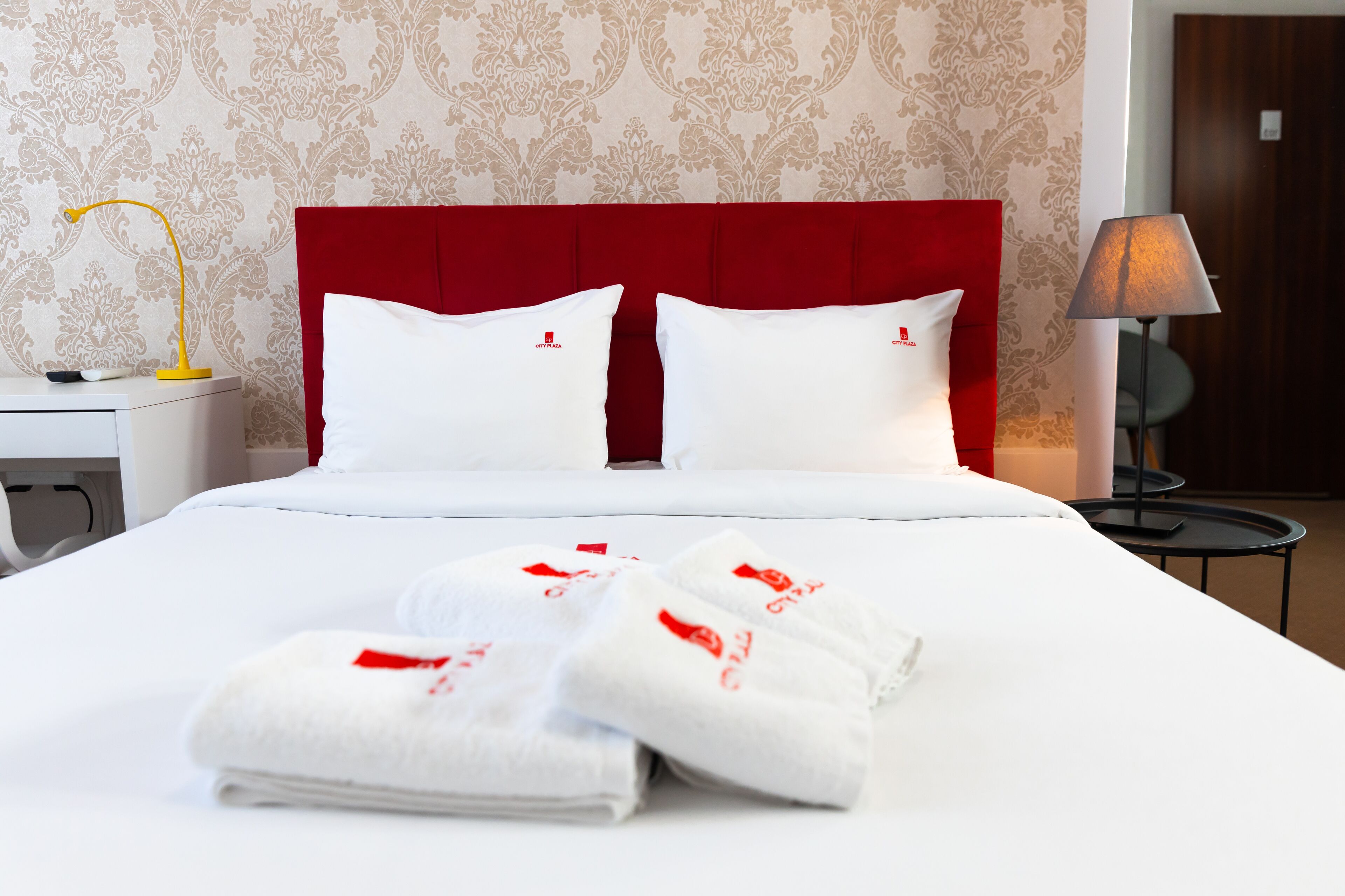 A hotel room with a red headboard, white bedding, and a neatly folded white towel with a red logo on the bed.