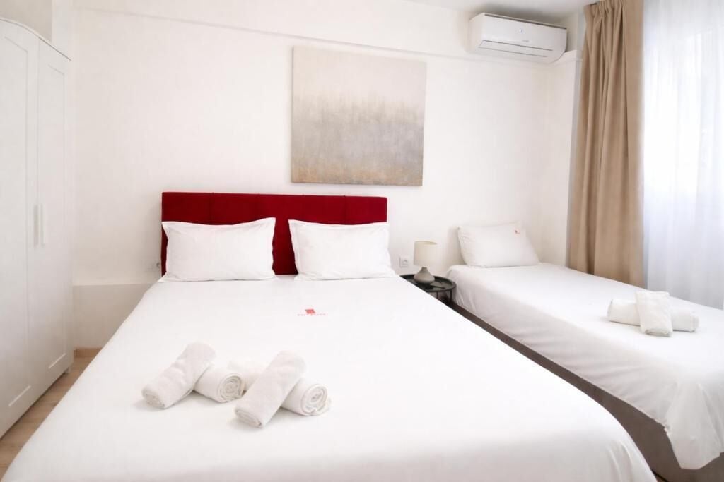 A hotel room with a large bed, two smaller beds, white linens, a red headboard, a painting on the wall, and an air conditioning unit.