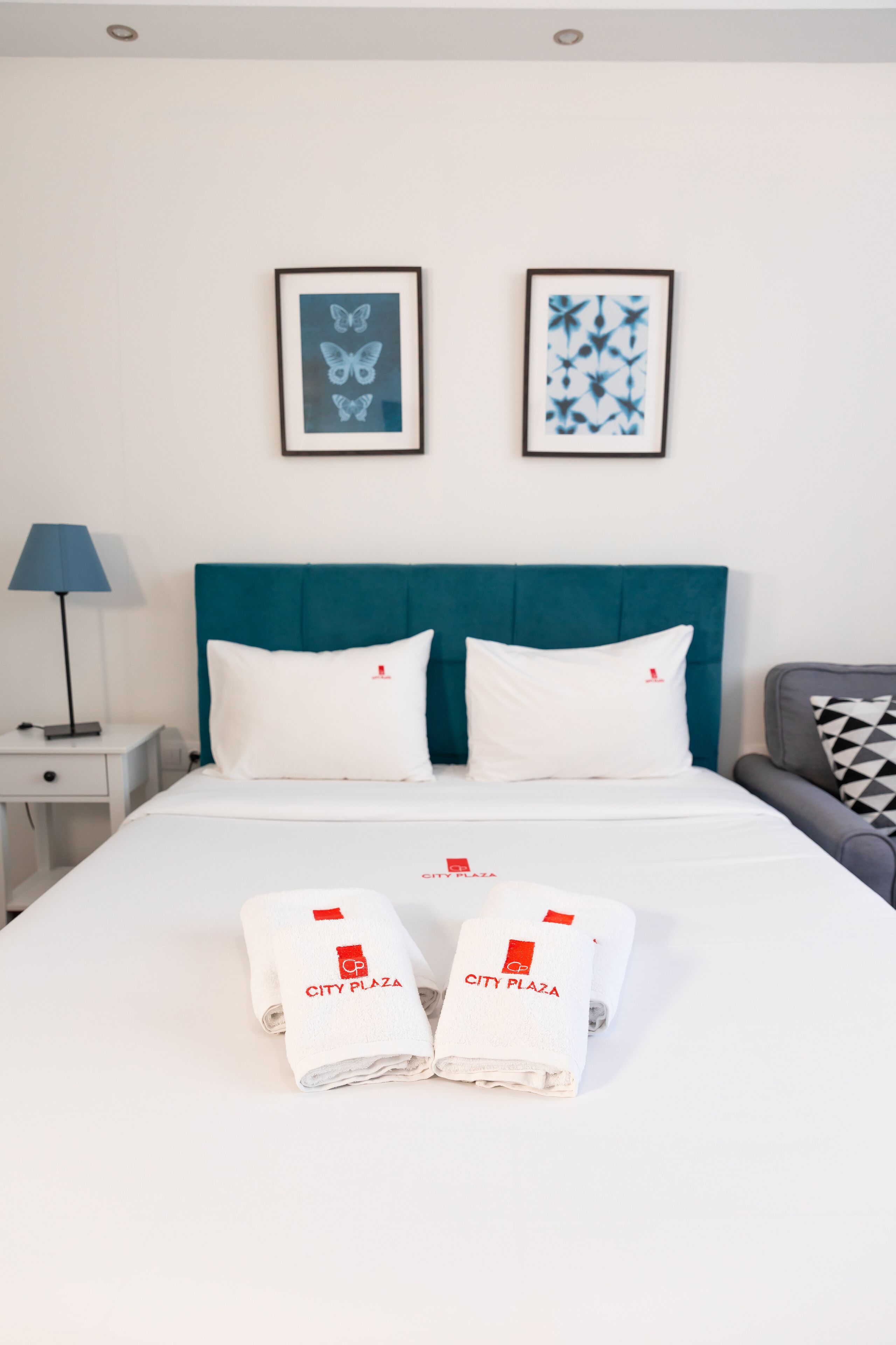 A hotel room with a teal headboard, white bedding, two framed butterfly artworks, a lamp, and two towels with "CITY PLAZA" branding.