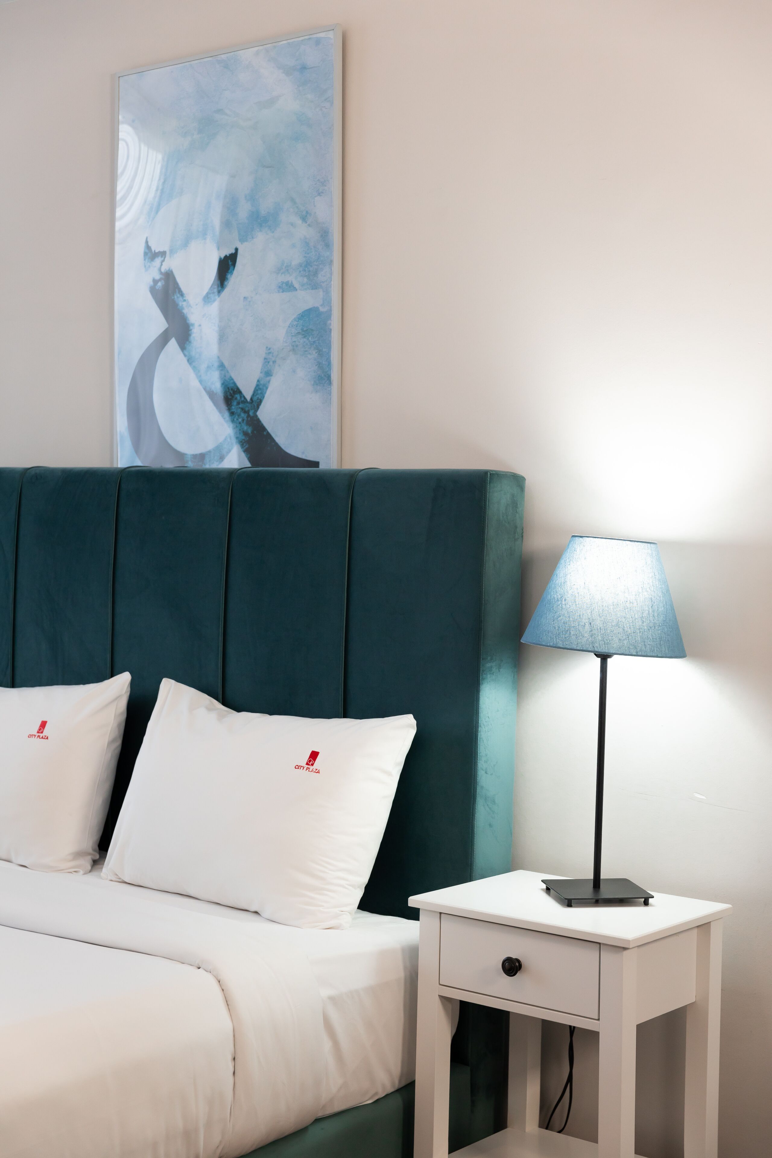 Una cama bien hecha con ropa de cama y almohadas blancas, un cabecero color aguamarina, una mesita de noche blanca con una lámpara y una obra de arte abstracta en la pared.