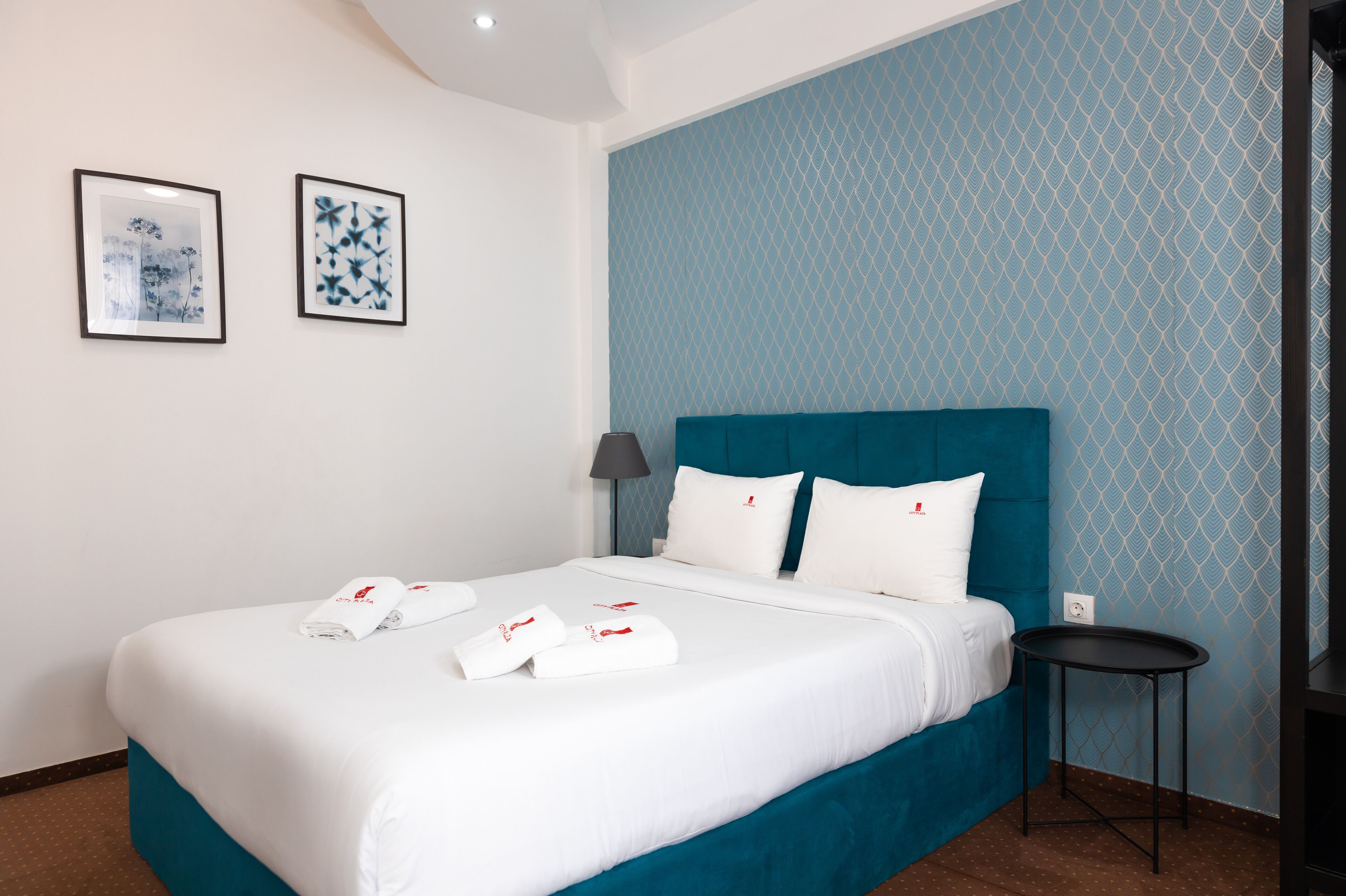 Een net opgemaakt bed met witte lakens en blauwgroene beddengoed, twee ingelijste foto's aan de muur en een klein zwart tafeltje ernaast.