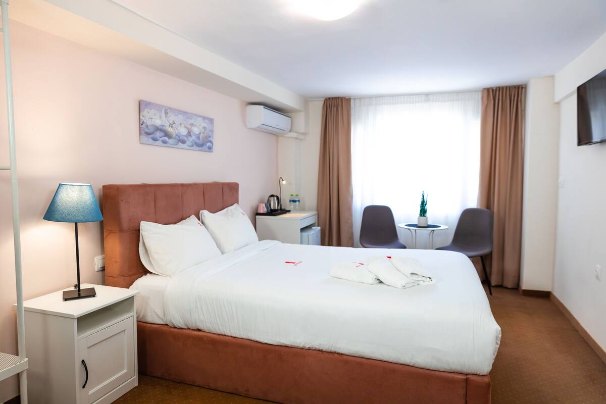 Een hotelkamer met een bed, nachtkastje, lamp, bureau, stoel en een televisie aan de muur.