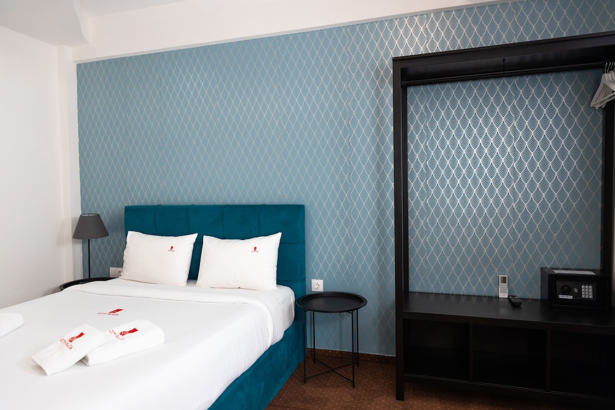 Een hotelkamer met een bed, een kleintje tafeltje, een stoel, een bureau en een spiegel.