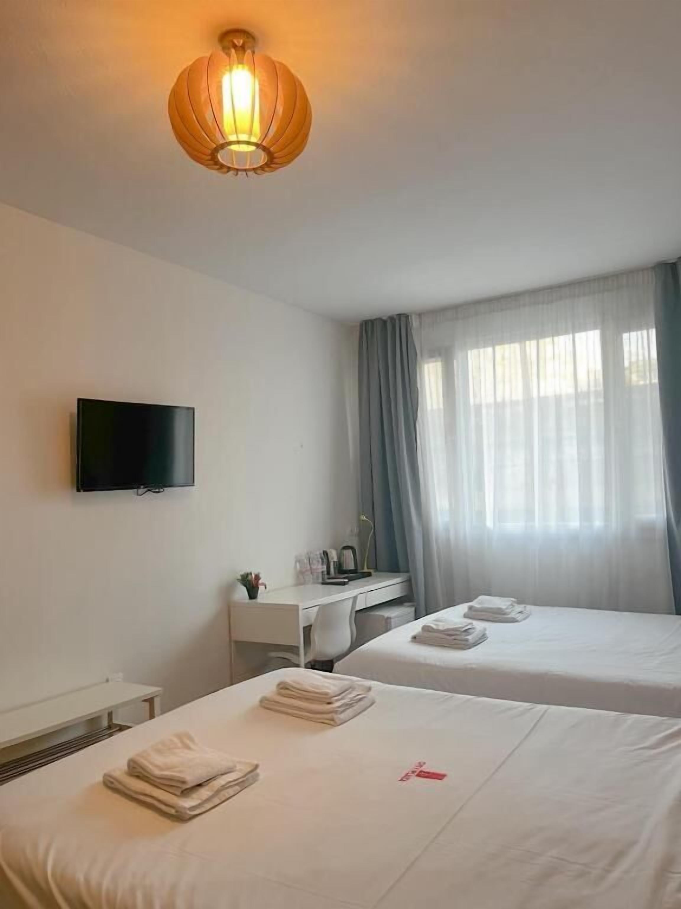 Ein Hotelzimmer mit zwei Betten, einem Schreibtisch, einem Fernseher und einem großen Fenster mit Vorhängen.