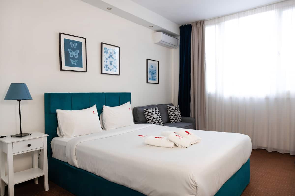 Een hotelkamer met een blauwgroen bed, witte beddengoed, een nachtkastje met een lamp en drie kunstwerken in lijsten.
