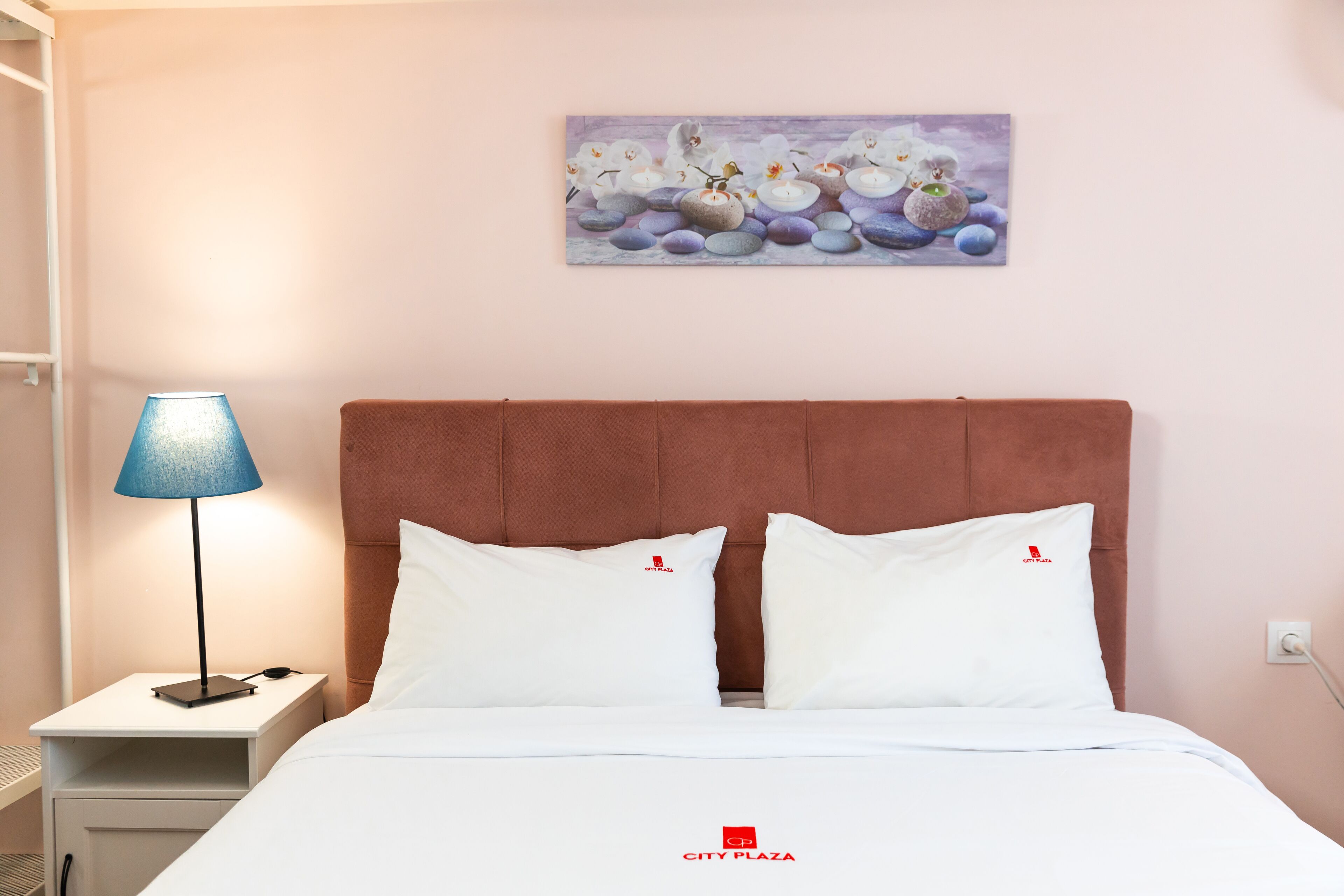 A hotel room with a bed, white pillows, a nightstand with a lamp, and a painting on the wall.