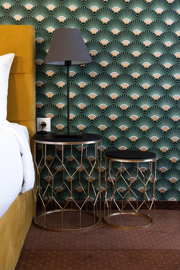 Une pièce avec un papier peint à motifs, un lit recouvert d’une couette blanche, une lampe et deux petites tables rondes à structure métallique.