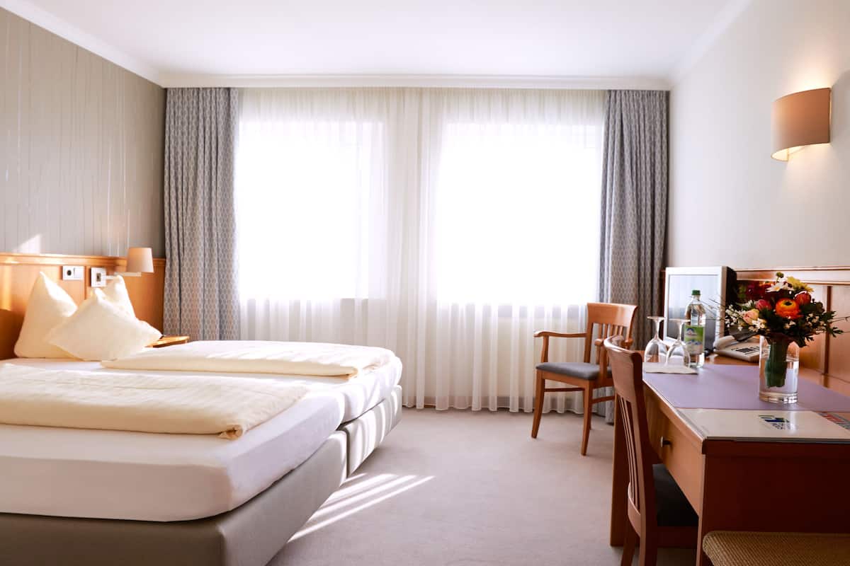 A hotel room with two beds, a desk with a chair, a vase of flowers, and a large window with curtains.