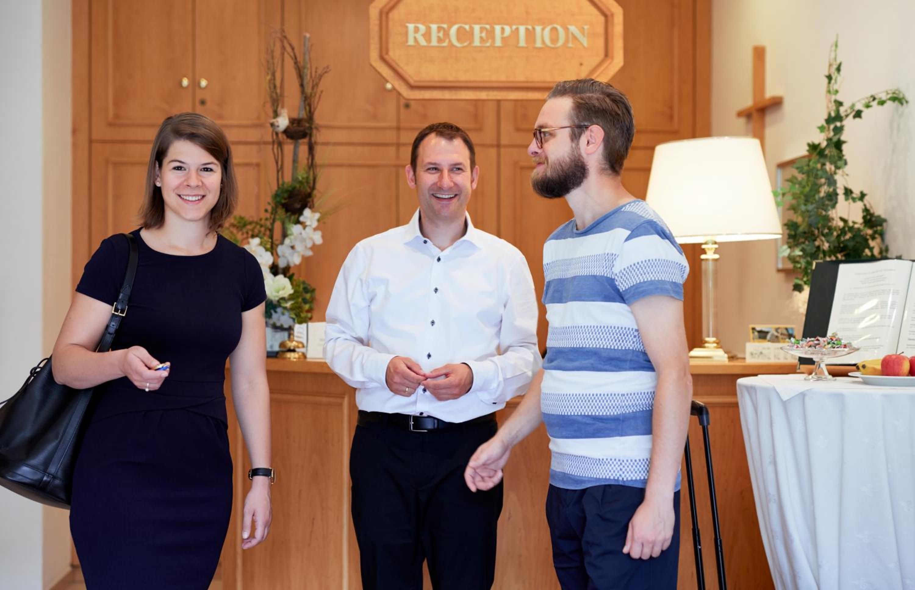 Three people standing in a reception area with a wooden reception desk, a sign that reads 'RECEPTION', and a table with a lamp and some items on it.