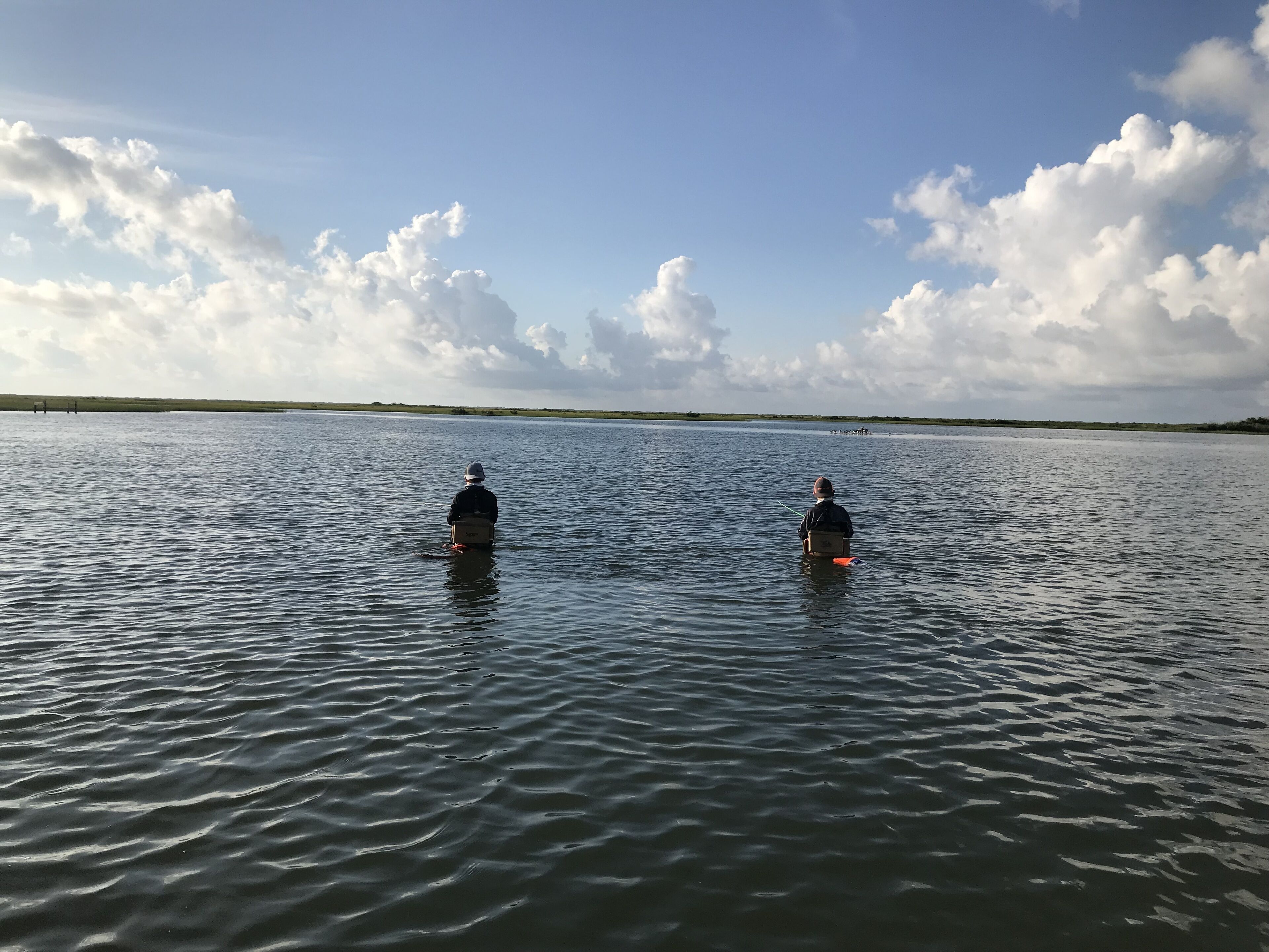 Two people sitting on boats in a calm body of water with a clear sky and clouds.
