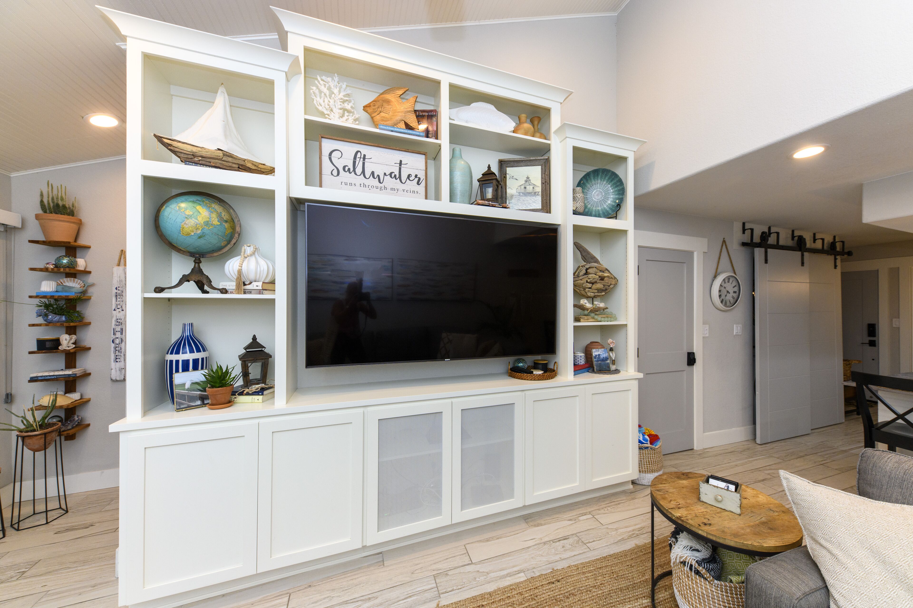 Ein Wohnzimmer mit einem großen Flachbildfernseher, der an einer weißen Unterhaltungsecke befestigt ist, umgeben von Regalen mit dekorativen Accessoires und Pflanzen.