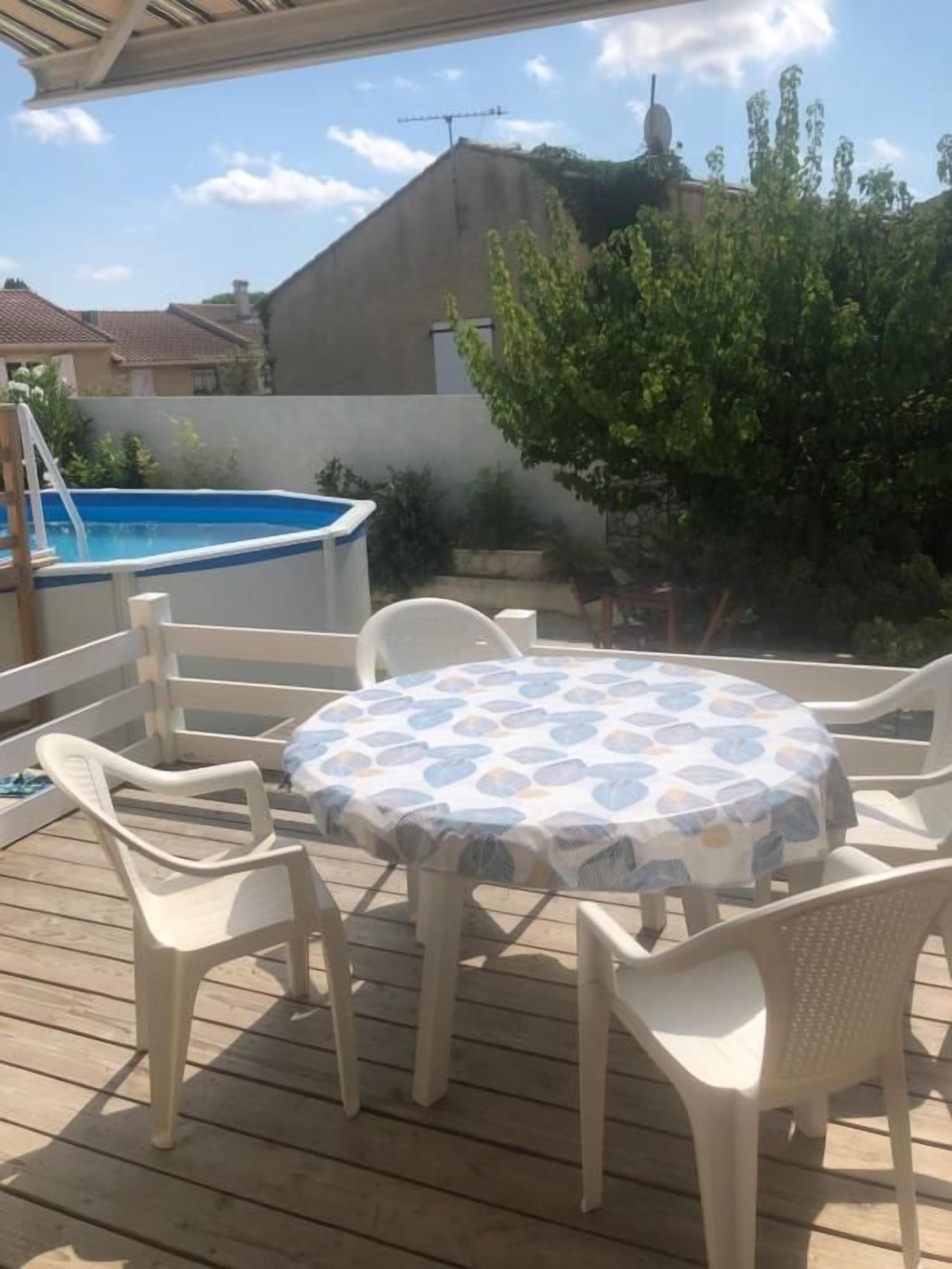 A wooden deck with a round table and chairs, a swimming pool, and a view of houses and trees.
