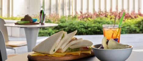 A sandwich on a wooden board, a glass of iced tea with a straw, and a bowl of chips on a table.