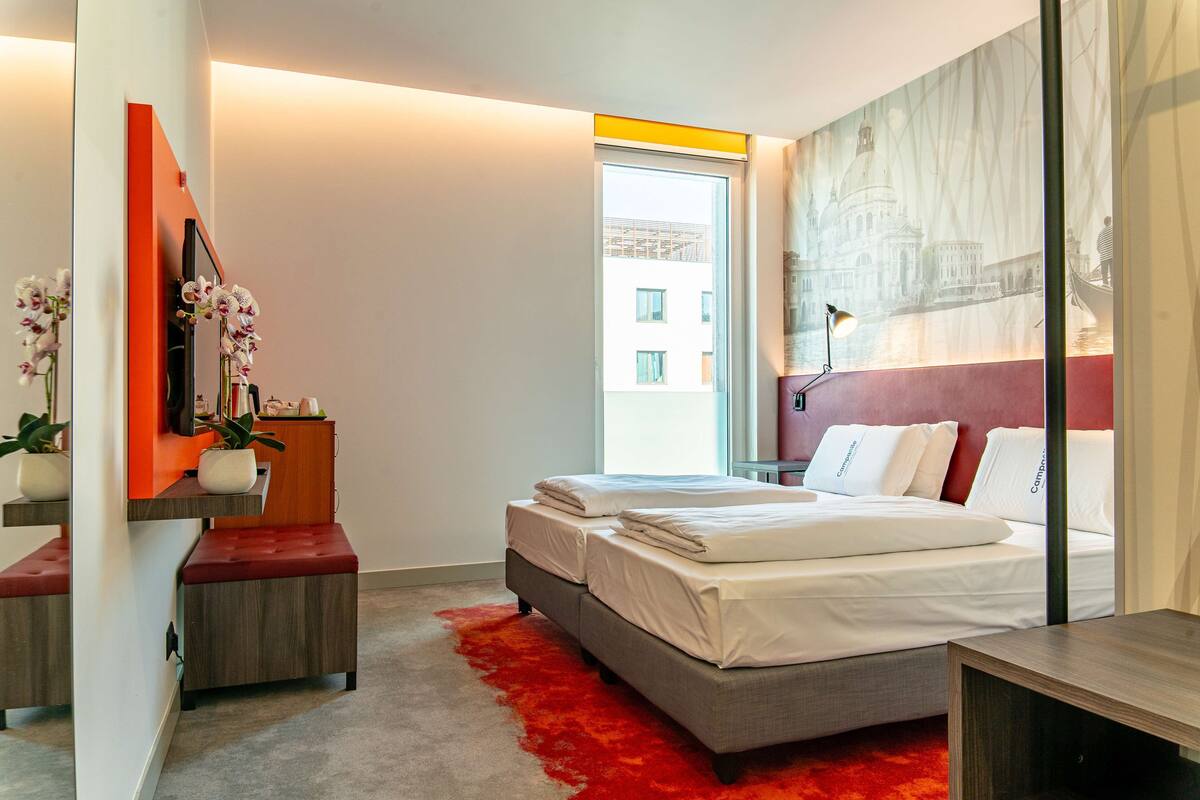 A modern bedroom with a large bed, a red carpet, a desk with a chair, a mirror, and a window with a view of buildings.