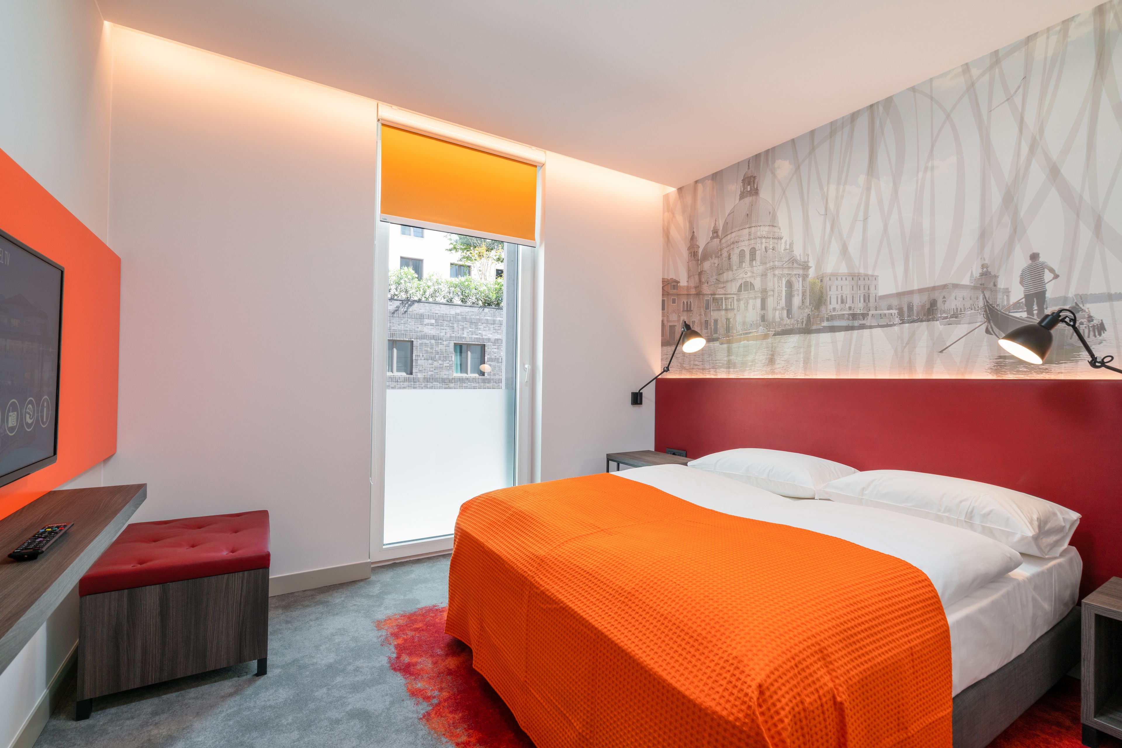 A modern hotel room with a large bed, a red accent wall, a cityscape mural, a desk with a remote control, and a red carpet.