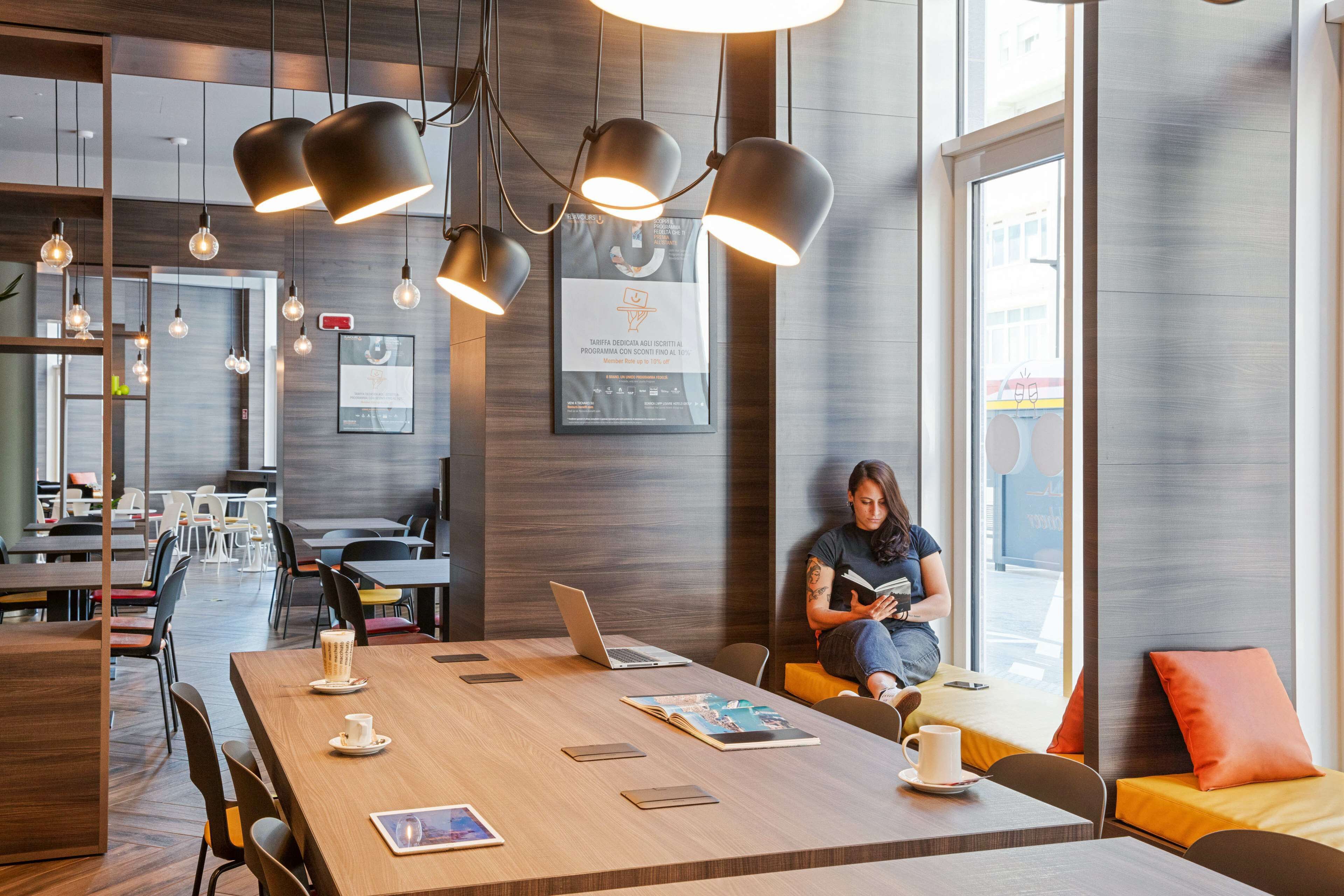 Spazio ufficio moderno con un lungo tavolo di legno, diverse sedie e un portatile. Sono presenti luci a sospensione, un certificato incorniciato appeso al muro e una persona seduta su un divano che legge un libro.