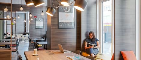 Un espacio de oficina moderno con una larga mesa de madera, varias sillas y un ordenador portátil. Hay luces colgantes, un diploma enmarcado en la pared y una persona sentada en un sofá leyendo un libro.