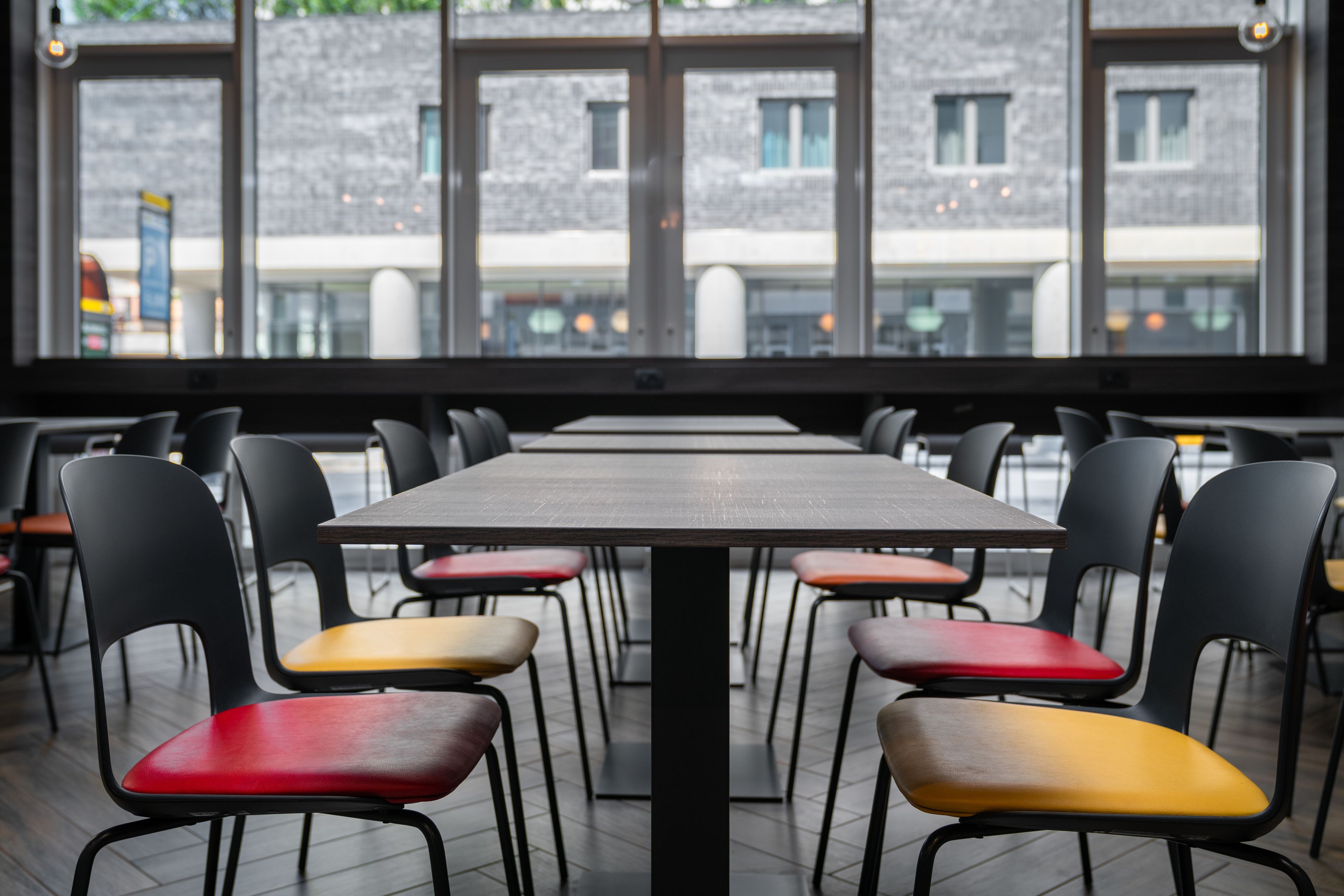 A restaurant with a long table and multiple chairs, large windows with a view of buildings outside.