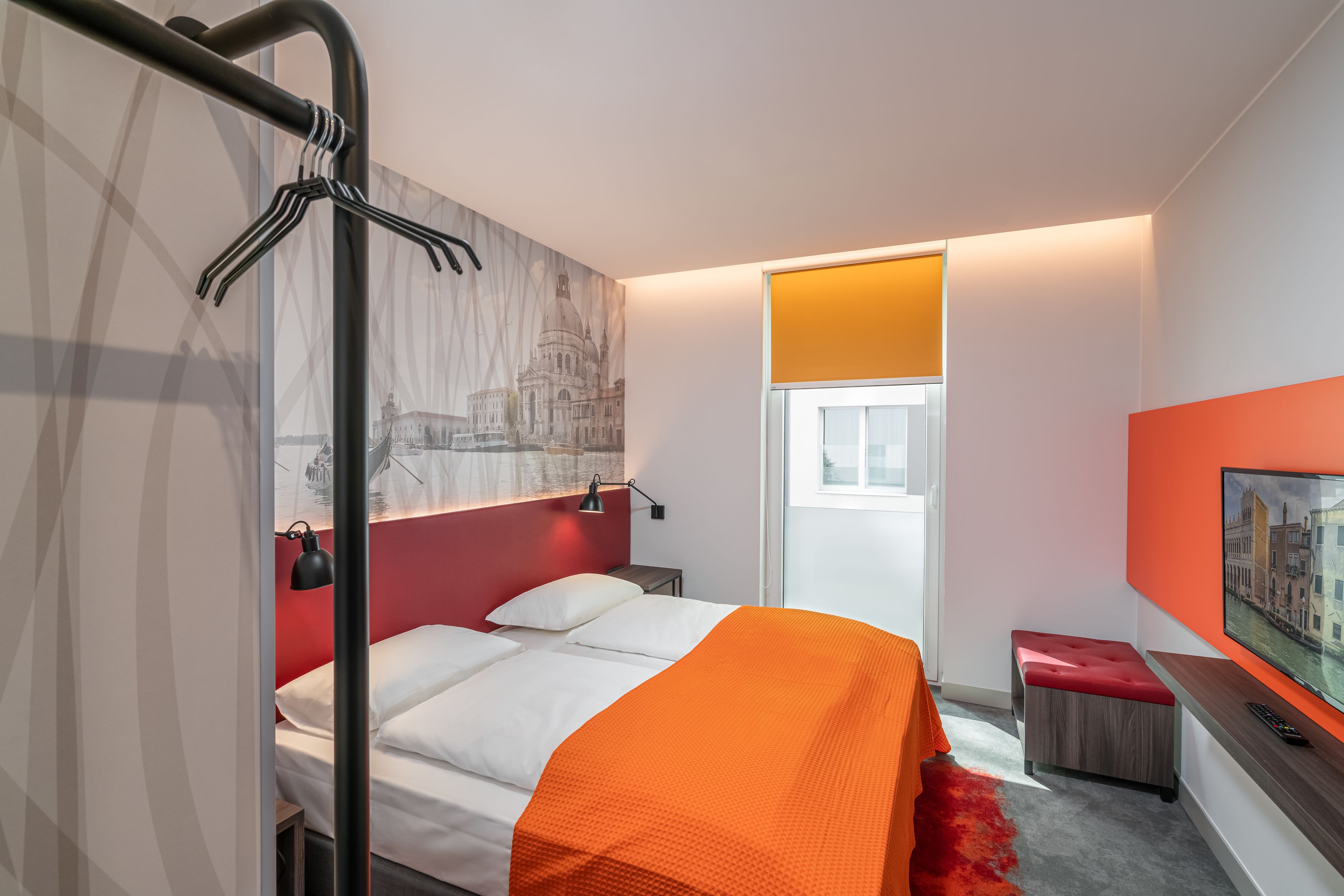 A modern hotel room with a bed, a red accent wall, a window with a view, a TV, and a red carpet.
