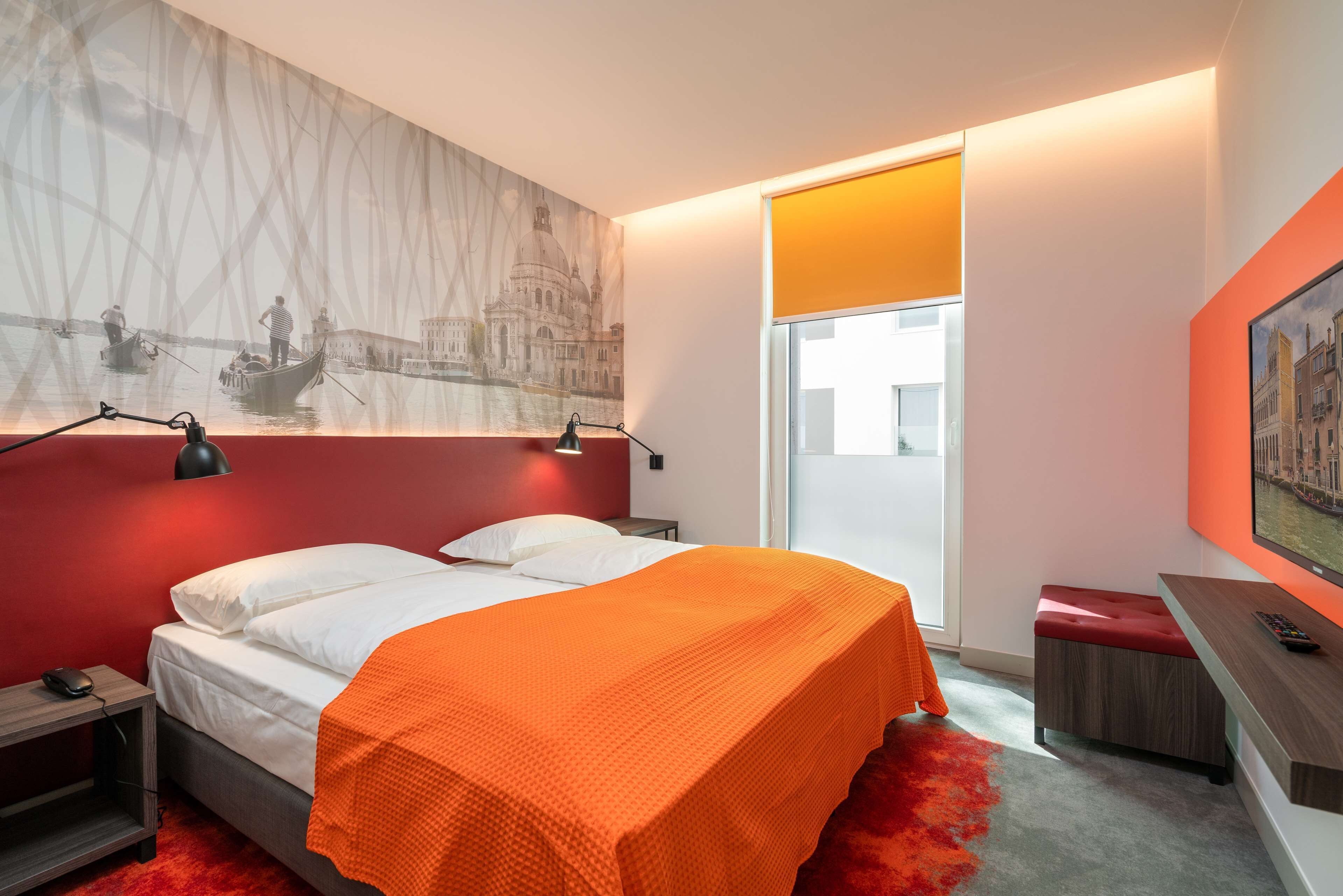 A modern hotel room with a bed, a desk, a chair, and a large window.