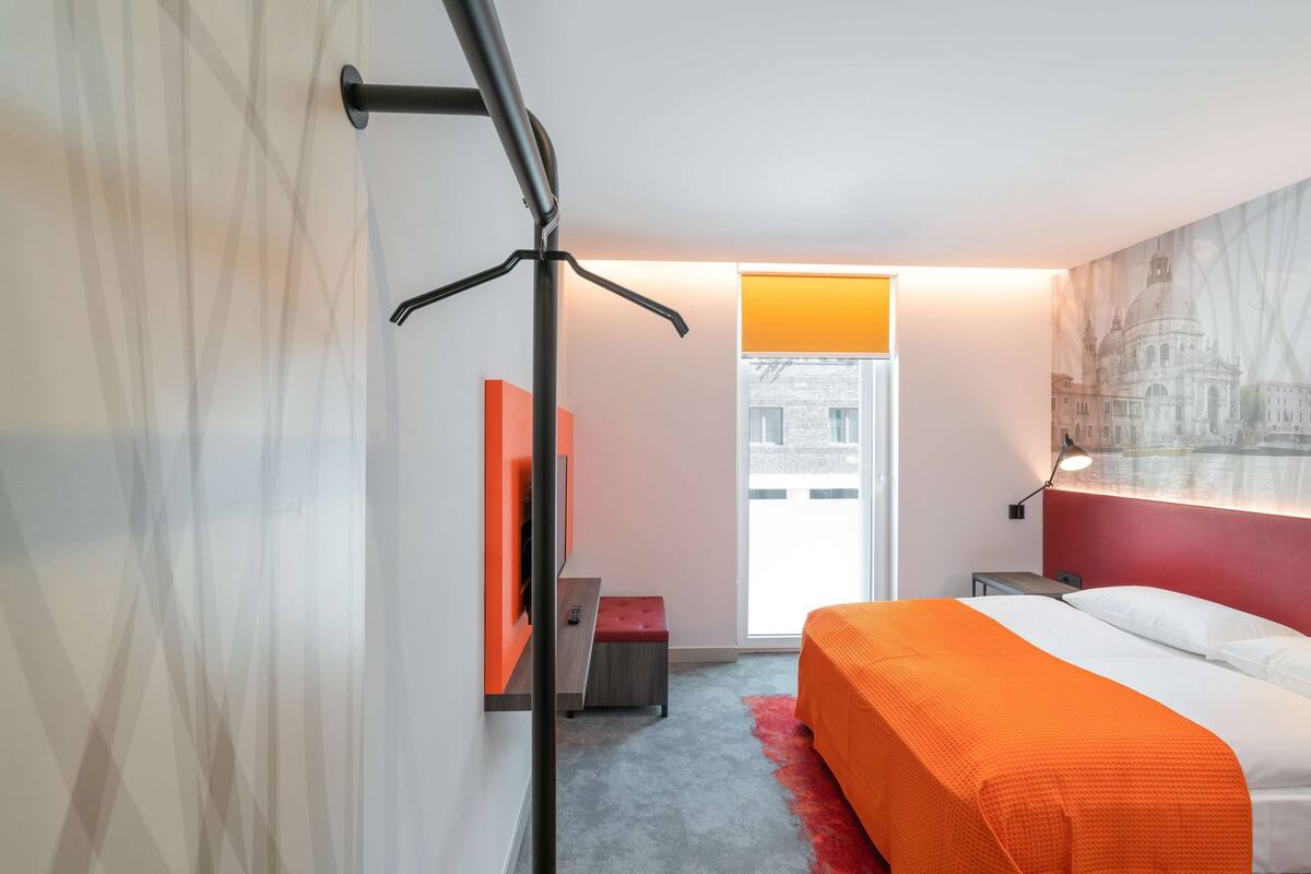 A modern hotel room with a large bed, a red accent wall, a cityscape view, and a red carpet.