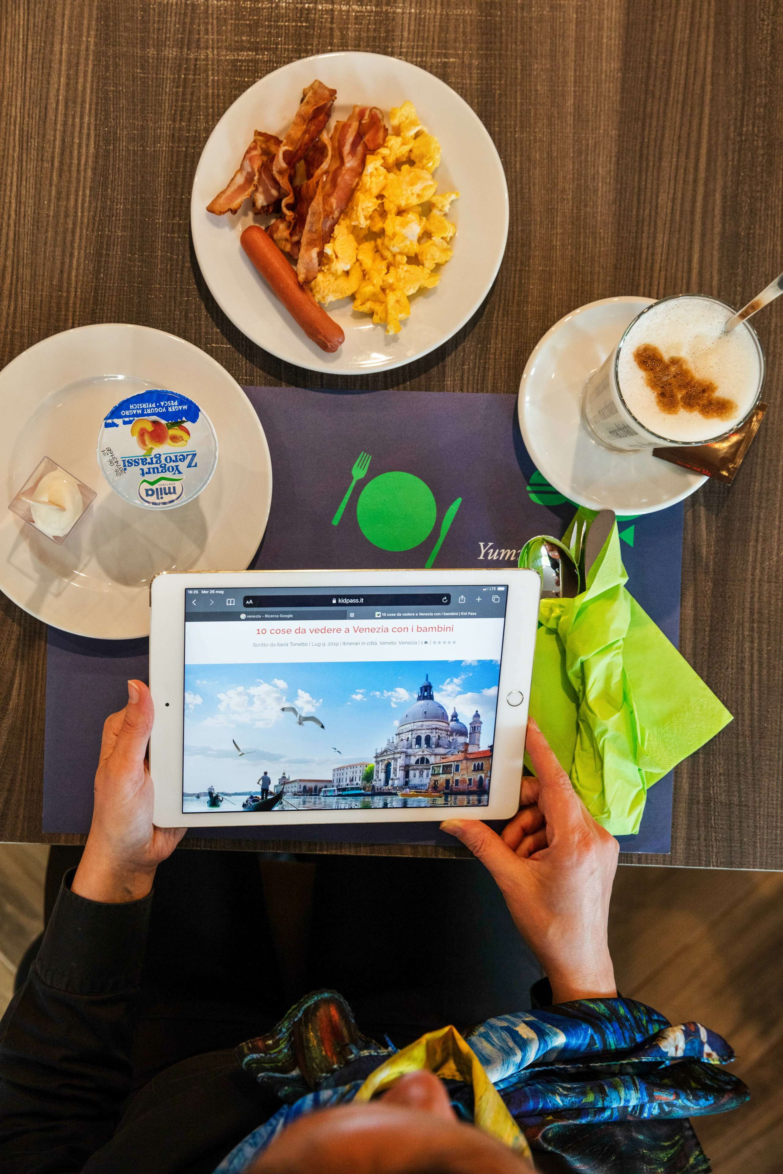 Une tablette affichant un site web de voyages, une assiette d’œufs brouillés et de bacon, un hot-dog, un verre de café et un sac cadeau vert, le tout posé sur une table.