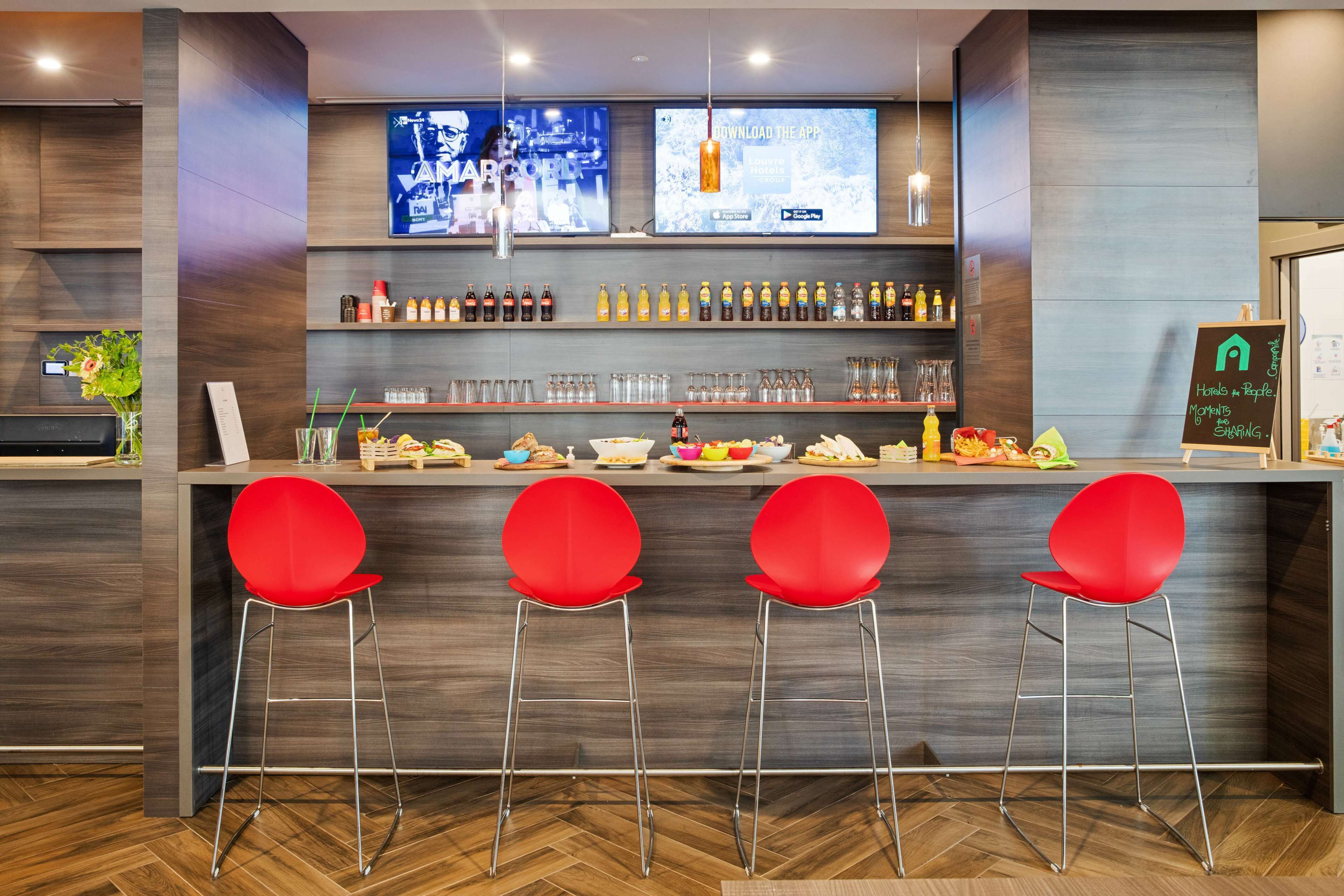 A modern bar area with a wooden counter, red bar stools, and a variety of beverages and snacks.
