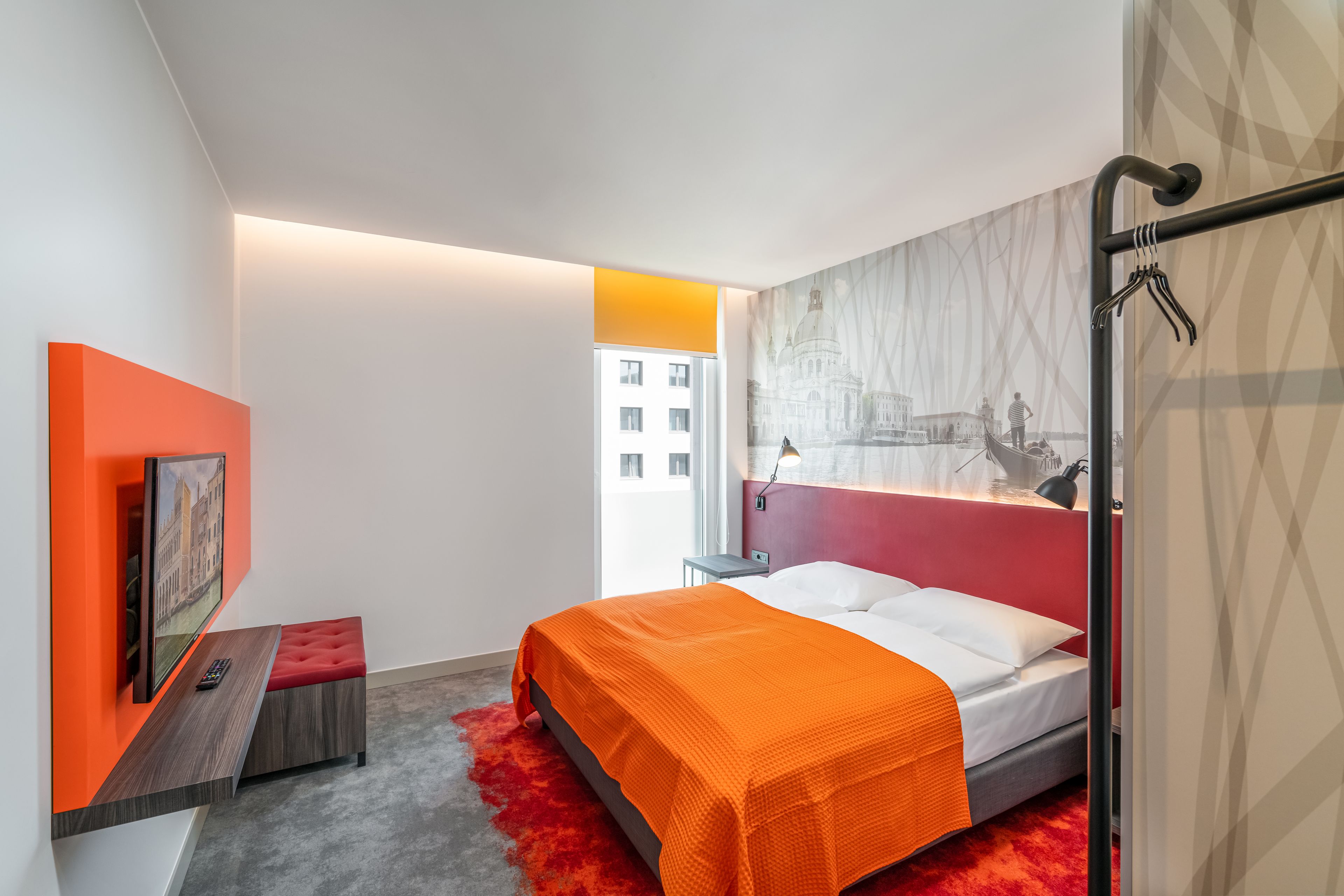 Une chambre d’hôtel moderne avec un lit, un mur d’accent rouge, un téléviseur fixé au mur et une vue sur le paysage urbain.
