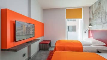 Une chambre d’hôtel avec un grand téléviseur fixé sur un mur orange, un lit recouvert d’une courtepointe orange, un petit pouf rouge et un tableau représentant un bâtiment accroché au mur.