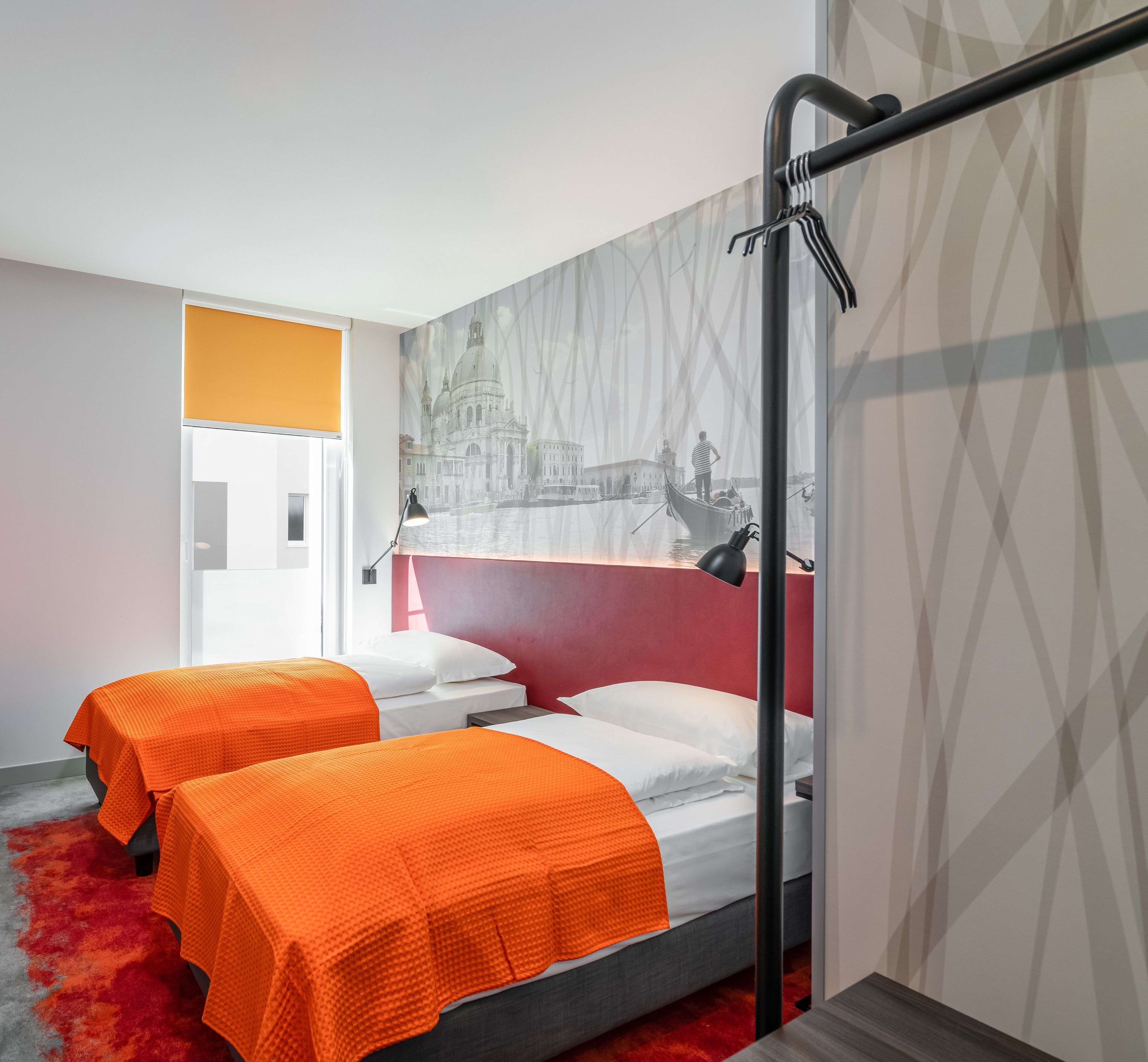 A modern hotel room with two beds, a red accent wall, and a cityscape mural.