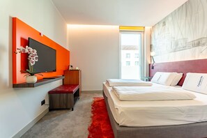 Une chambre d’hôtel moderne avec un grand lit, un téléviseur fixé au mur, un mur d’accent rouge et une vue sur le paysage urbain.