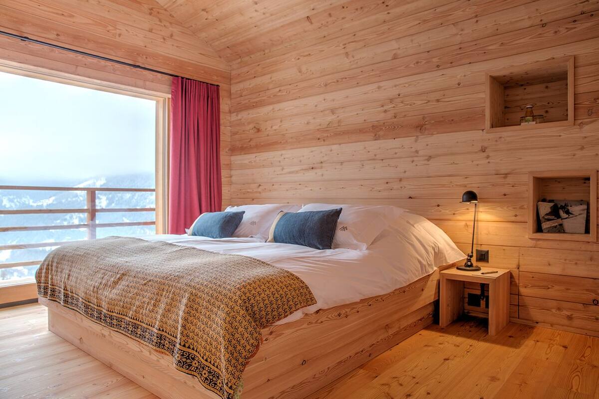 A wooden cabin bedroom with a bed, a nightstand, a lamp, and a view of the mountains.
