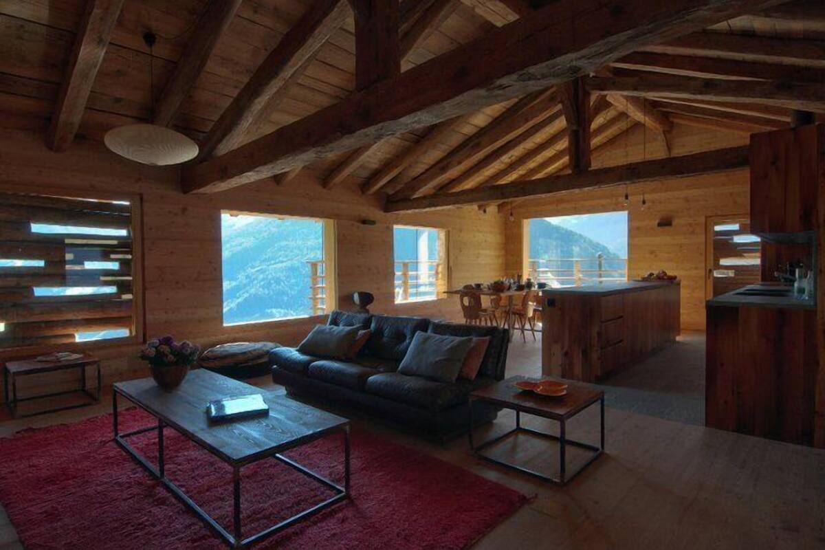 A spacious living area with a wooden ceiling, large windows offering mountain views, a dark-colored sofa, and a kitchen area.