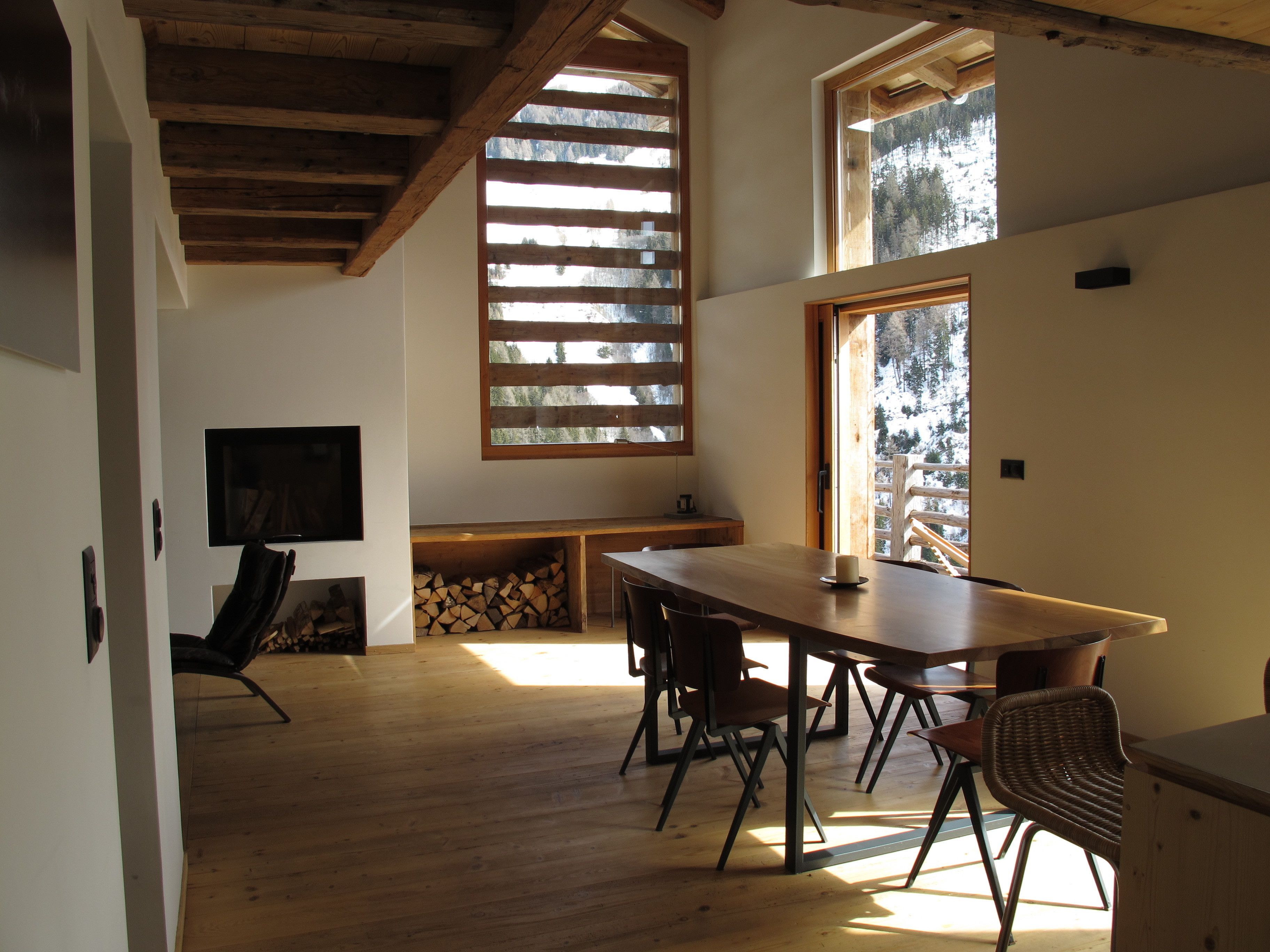 A spacious room with a large wooden table and chairs, a TV mounted on the wall, and a window with a view of snow-covered trees.