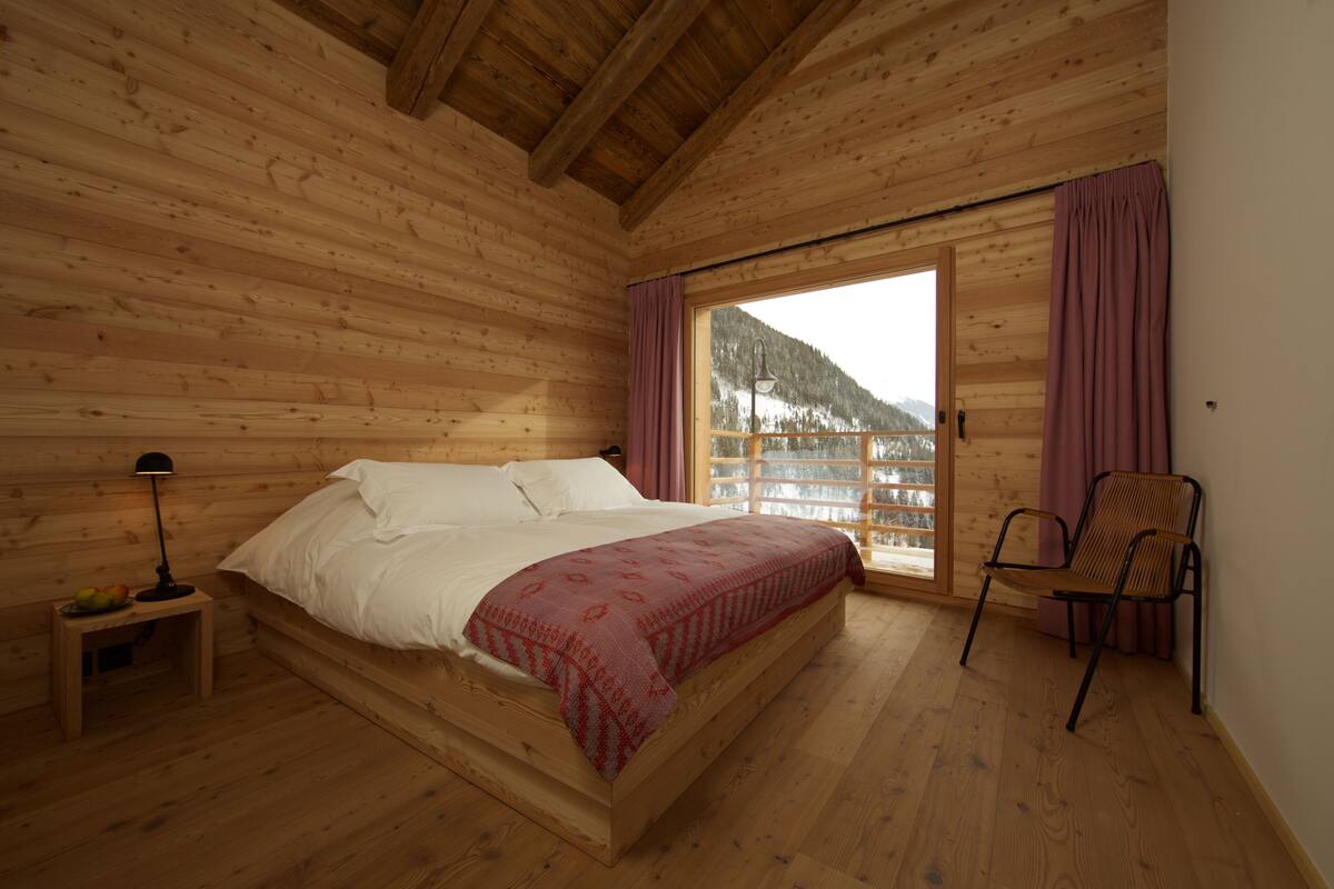 A wooden cabin bedroom with a bed, a chair, a nightstand, and a window with curtains.