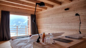 Ein Zimmer in einer Holzhütte mit einem Bett, zwei Schreibtischlampen, einem Fenster mit Vorhängen und Bergblick.