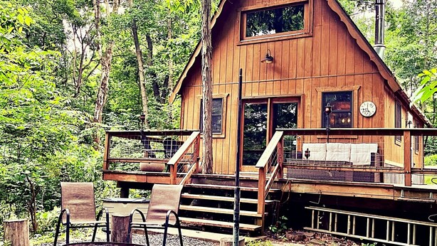 A wooden cabin with a porch, a fire pit, and a view of the surrounding forest.