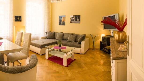A living room with a sofa, a glass coffee table, a dining table with chairs, a chandelier, and a TV on a wooden cabinet. - Apartment Annabelle (Vienna)