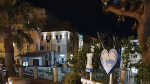 Vista nocturna de un letrero con la palabra 'TROPEA' y un símbolo de corazón, rodeado de macetas y una zona de grava.