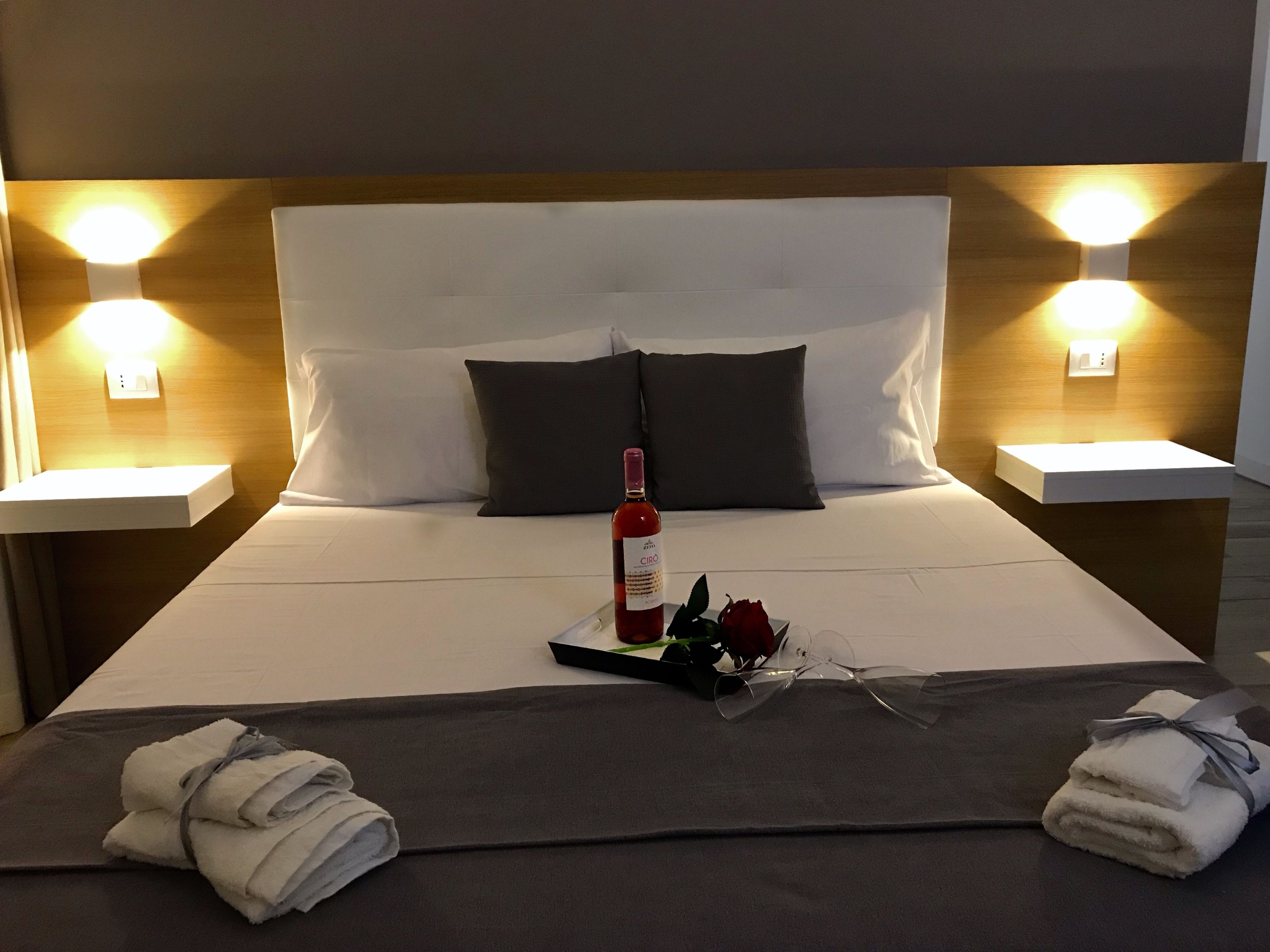 A neatly made bed with a bottle of wine, a rose, and glasses on a tray, towels folded at the foot of the bed, and two bedside tables with lamps.