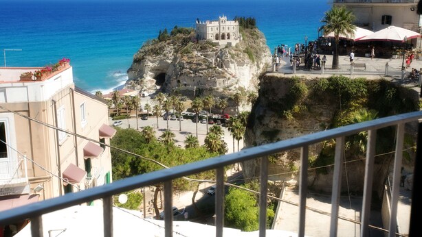 Vista costera con una estructura similar a un castillo sobre un acantilado, un área pavimentada con palmeras y un edificio con balcones.