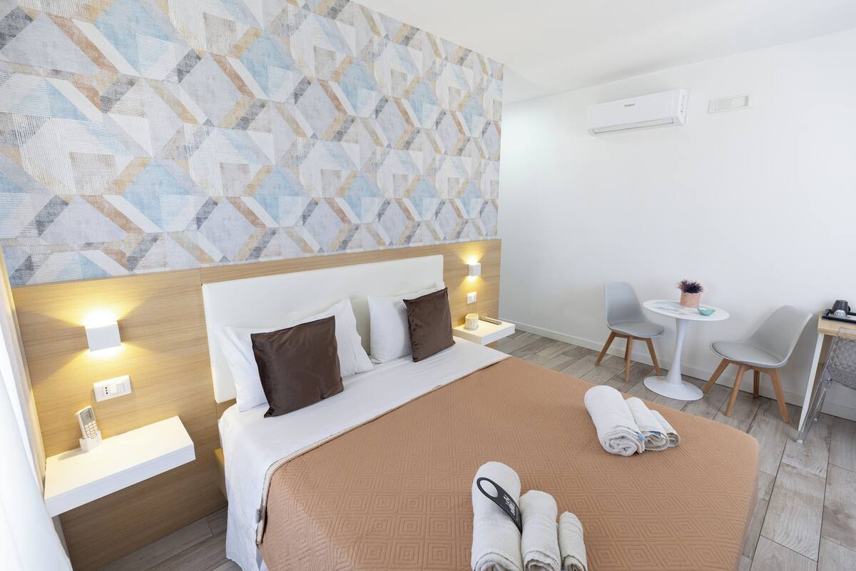 Una habitación de hotel con una cama grande, una mesita de noche, un escritorio con silla, una pequeña mesa redonda con una silla y un aire acondicionado de pared.