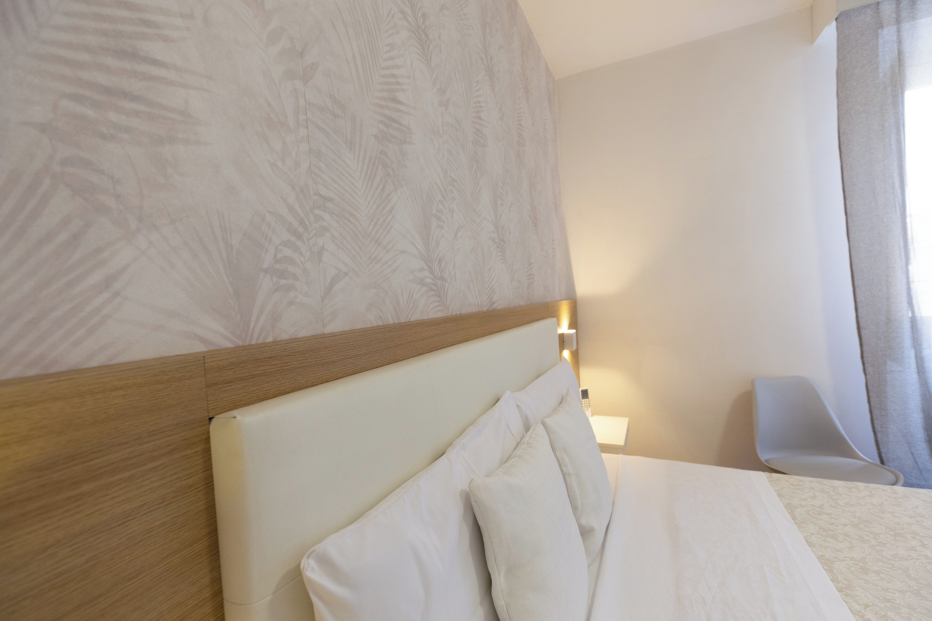 Una habitación de hotel con cabecera de madera, ropa de cama blanca, una mesita de noche con lámpara y vistas a una ventana con cortinas.