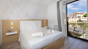 Habitación de hotel con una cama grande, una mesita de noche, un estante en la pared y vistas a la calle desde un balcón.
