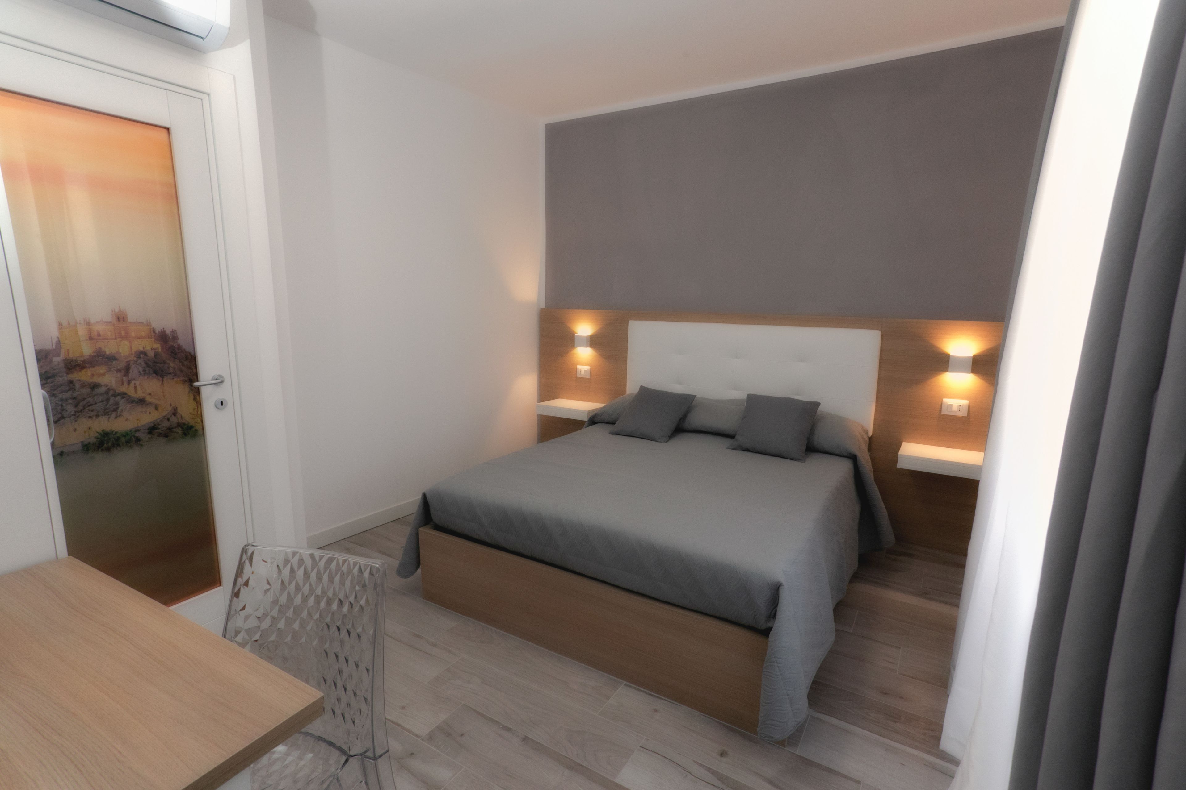 Un dormitorio moderno con una cama grande, dos mesitas de noche con lámparas, un cabecero de madera y vistas a un paisaje urbano a través de una puerta.