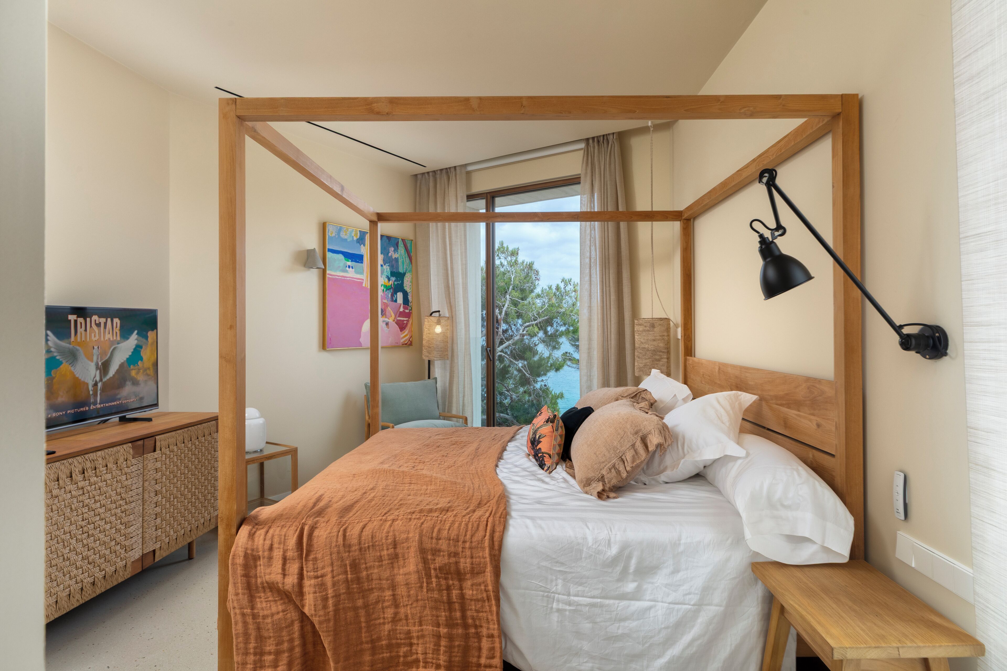 Een slaapkamer met een hemelbed, een houten hoofdbord, een nachtkastje, een bankje, een televisie op een kast, en een raam met gordijnen.