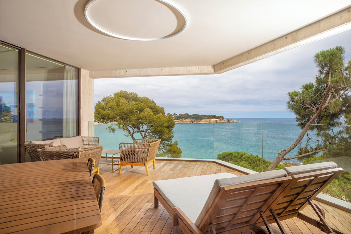 Een modern terras buiten met een houten tafel en stoelen, een glazen balustrade en uitzicht op zee en het land in de verte.